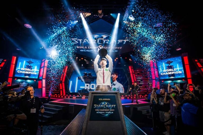 Esports player in a white jacket raises a trophy on a brightly lit stage with blue confetti during the StarCraft World Championship Series finals, crowd cheering in the foreground.