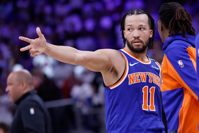 Basketball player in a blue Knicks jersey (#11) extending his arm and pointing during a game on the court and crowd in the background.