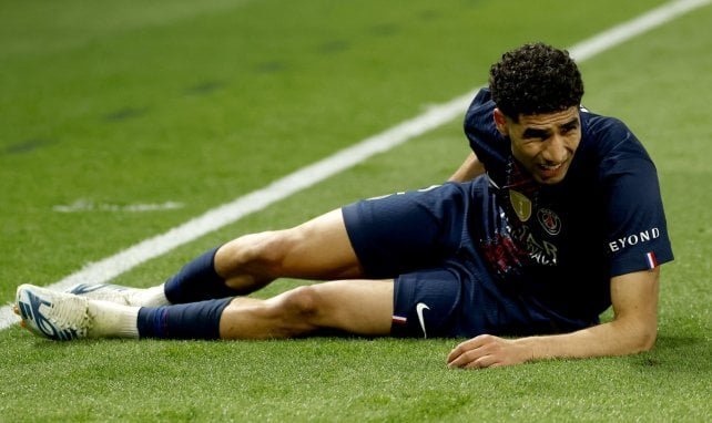 Soccer player in a dark blue kit lies on his side on a grass field near a white line, appearing injured during a match.