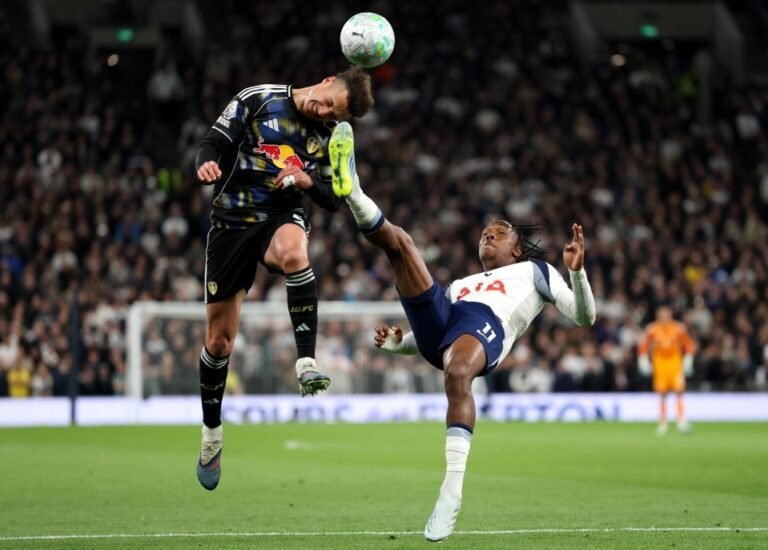 Two soccer players leap in the air competing for a header during a match, one in a dark kit with a Red Bull logo and the other in a white jersey marked USA on the chest.